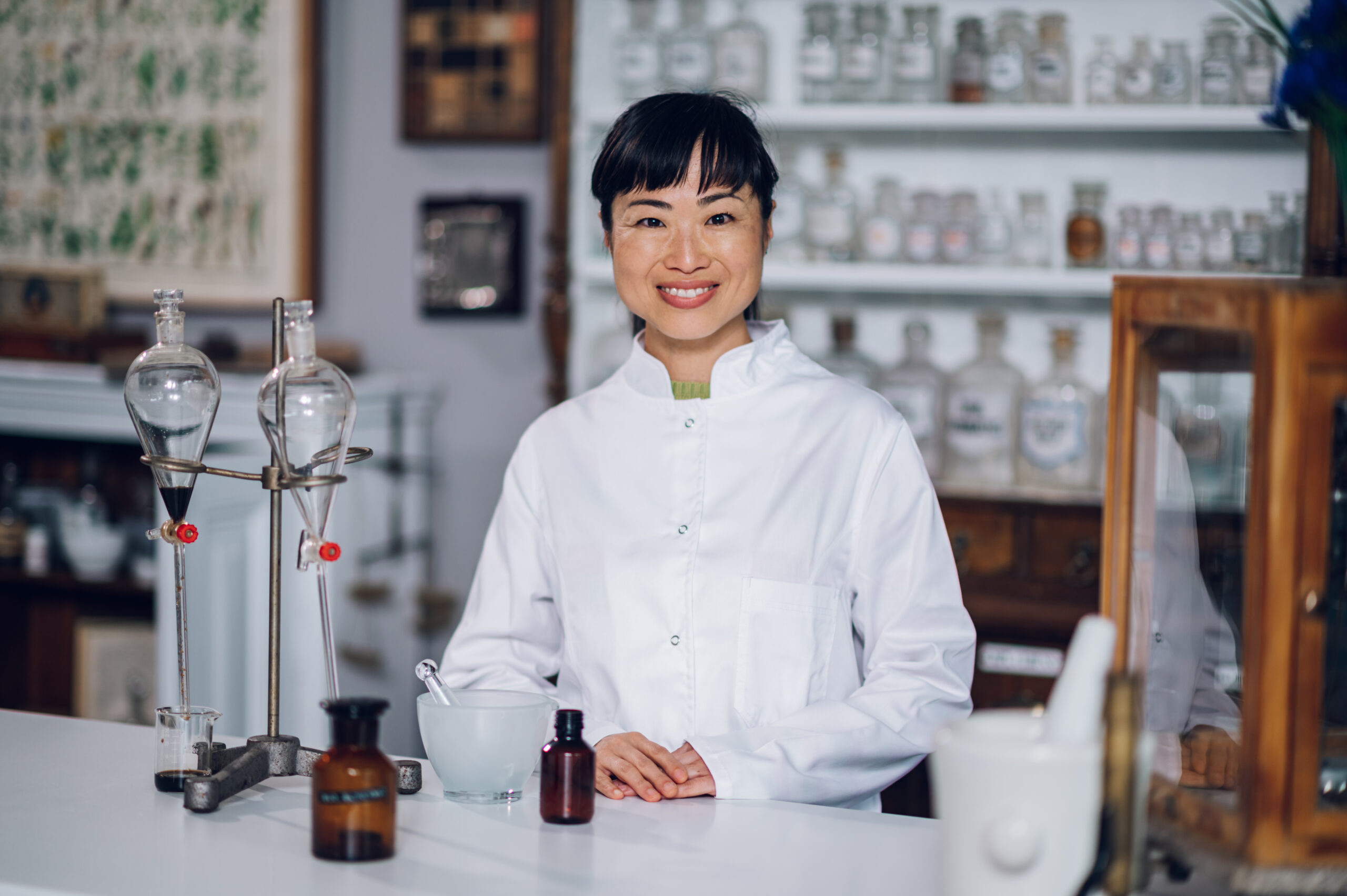 Portrait of a Japanese pharmacist in a lab coat standing with glass vessels and laboratory equipment and smiling at the camera. A happy technician is posing in a drugstore.