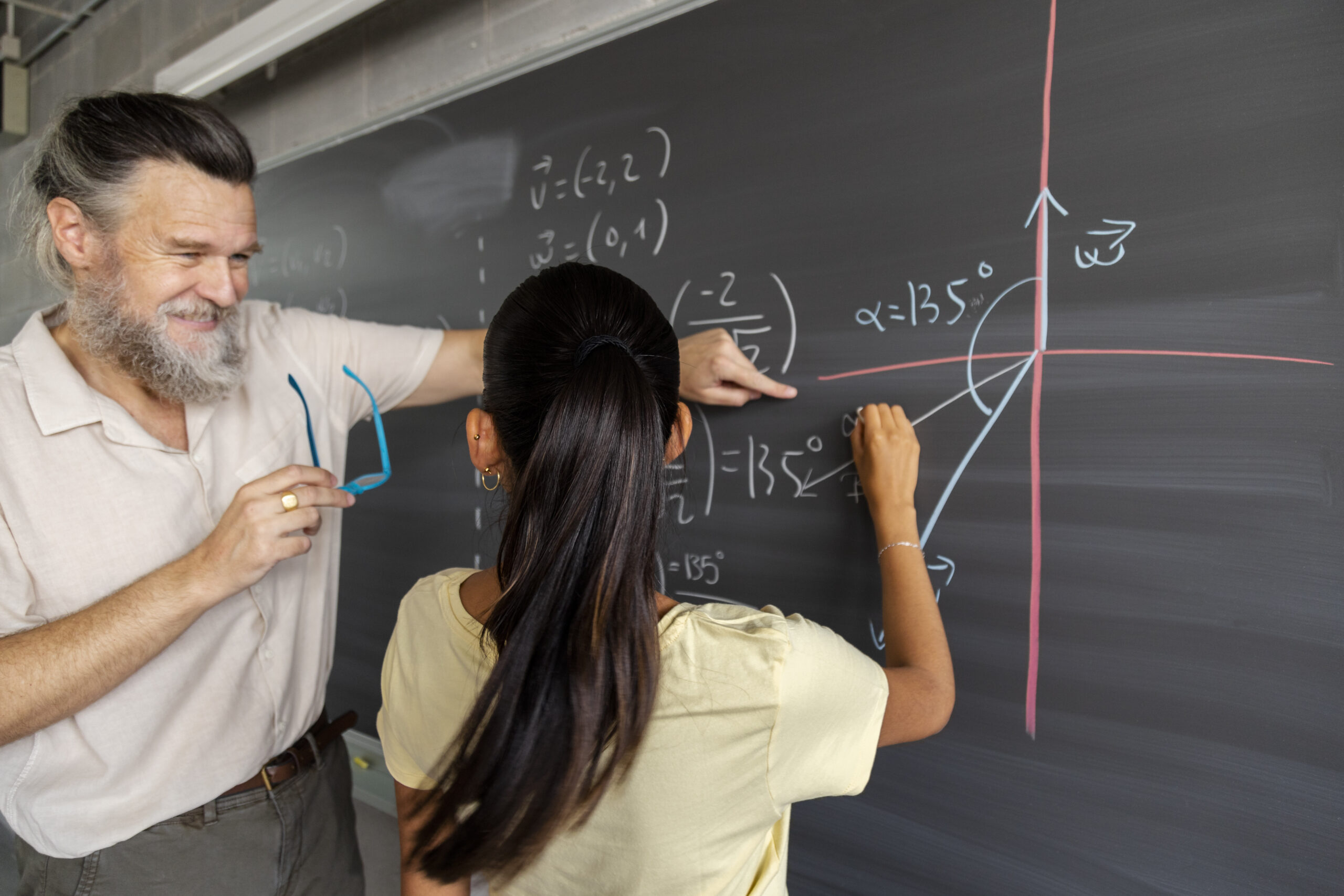 Smiling mature caucasian man teacher shows female teen high school student math problem resolution on blackboard. Education concept.