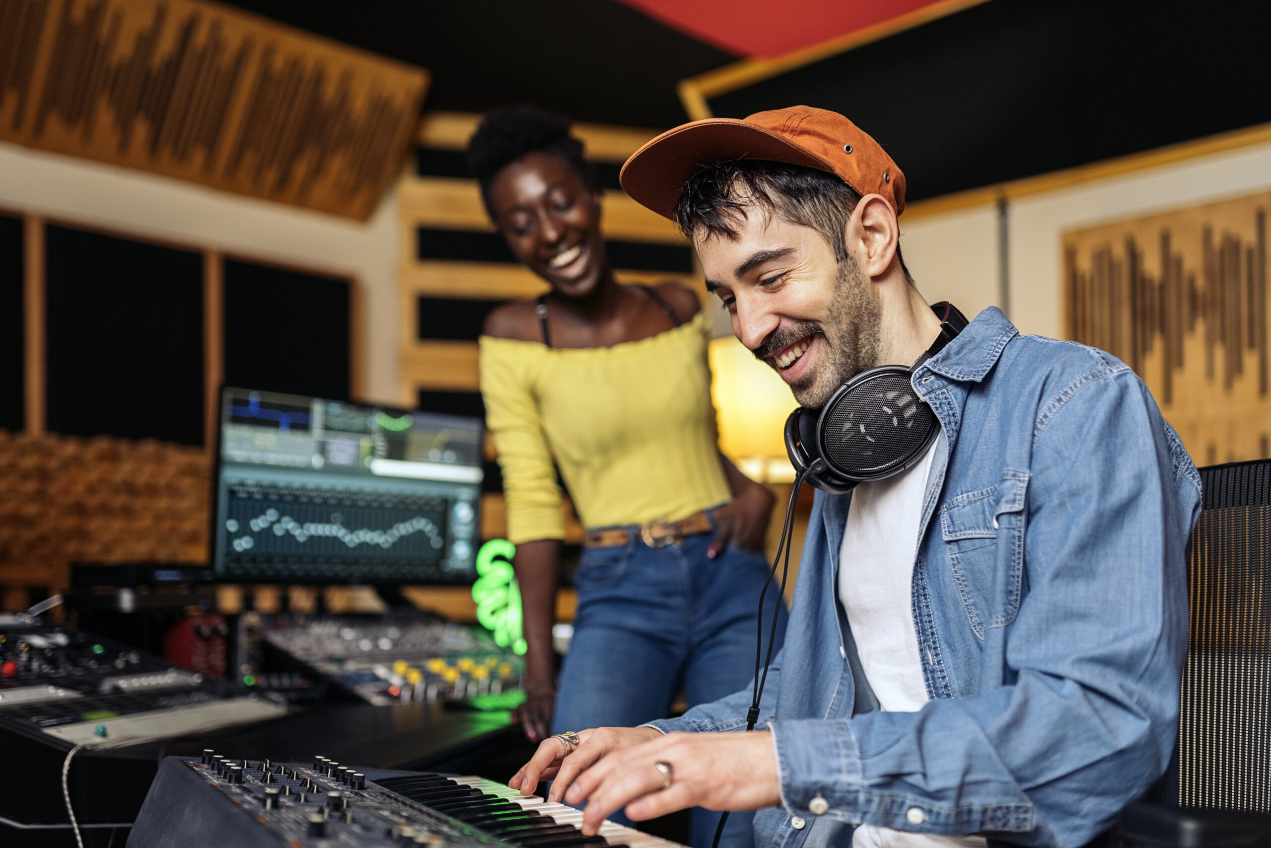 Stock photo of happy black woman in music studio and male producer using electronic piano keyboard.